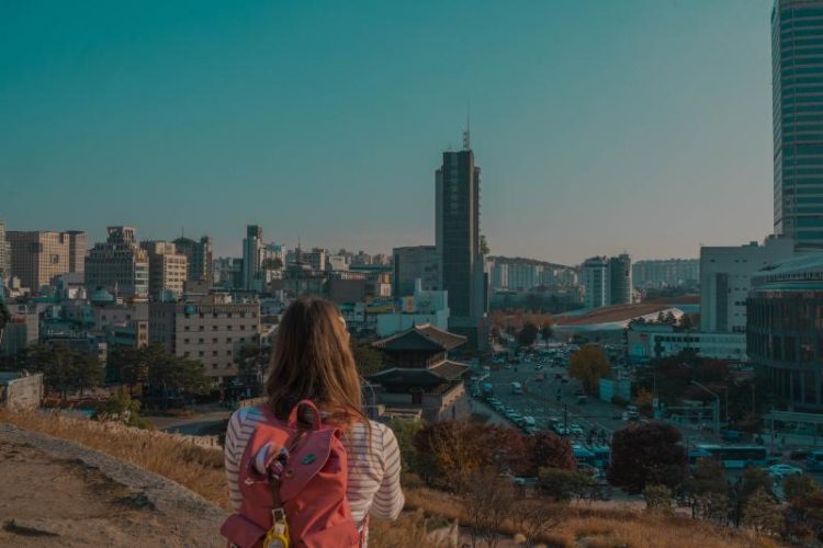 woman with pink backpack looks at city skyline with skyscrapers and historic city gate in Seoul