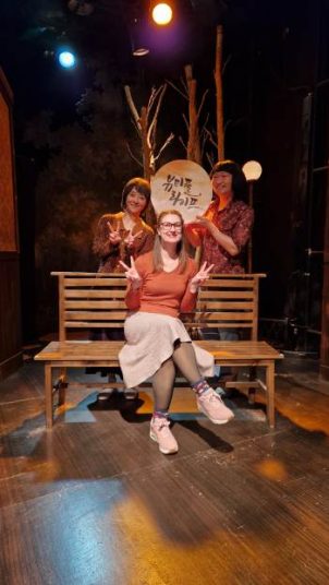 Annemarie sitting on a bench with two actors holding up signs behind her