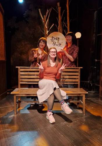 Annemarie sitting on a bench with two actors holding up signs behind her