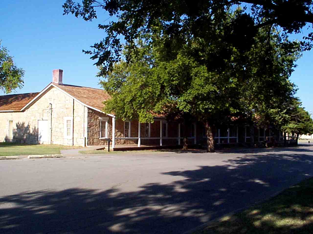 shady trees stand next to a paved road and a grey brick house